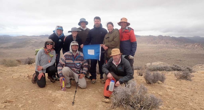 A group of people gather for a group photo amid a desert landscape. One of them holds a blue peter flag.